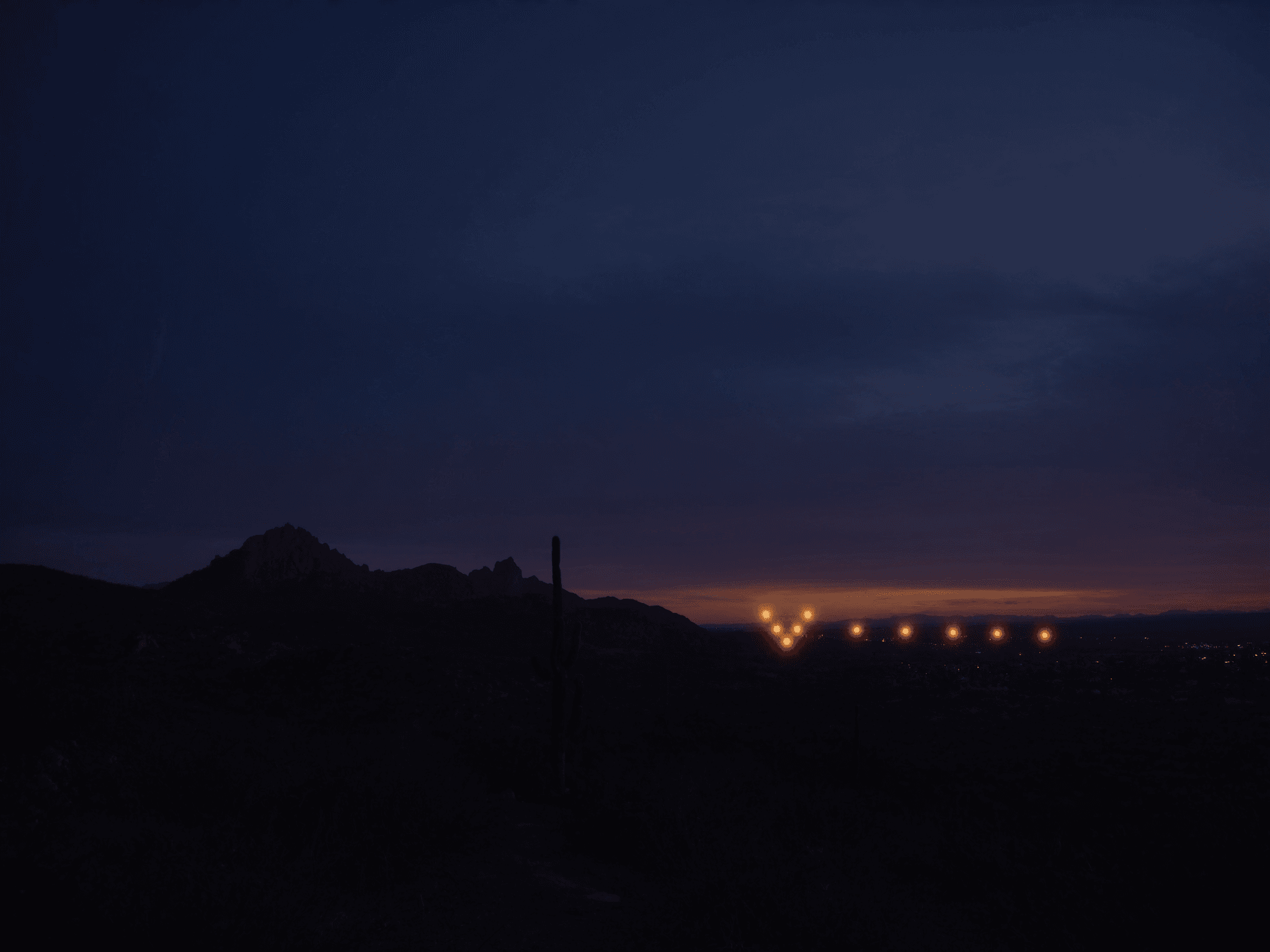 Sonoran desert at twilight with a faint V-formation of distant amber lights on the horizon