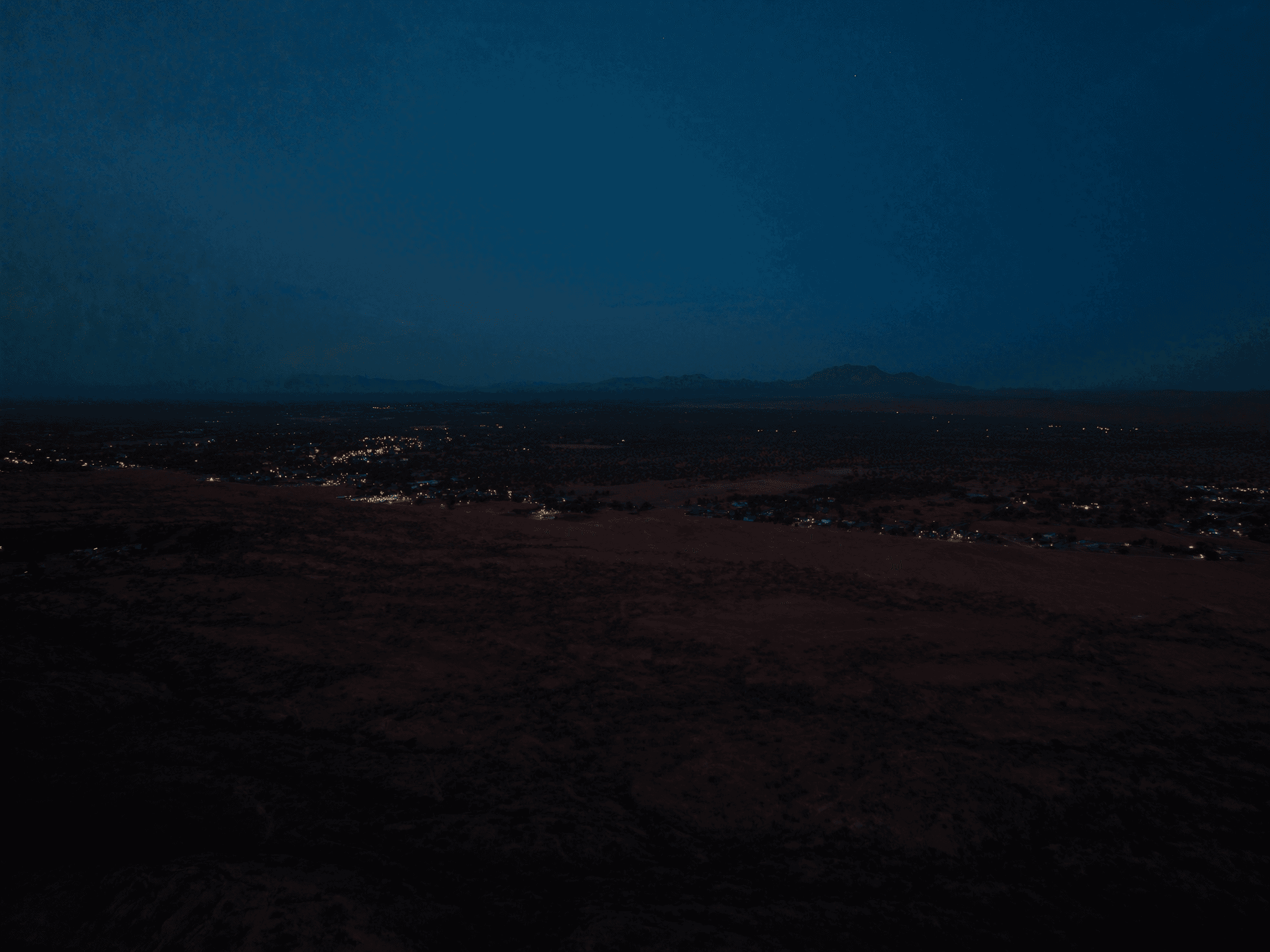 Aerial dusk view across a Southwest desert corridor with town lights and mountains